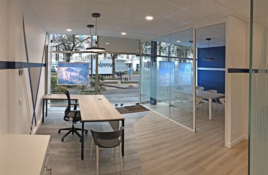 Bureau moderne avec de grandes fenêtres, deux bureaux et des chaises au premier plan, et une salle de réunion vitrée avec une table et des chaises à l'arrière-plan.