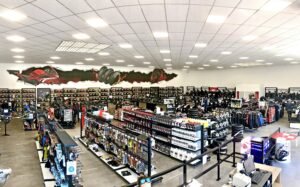Vue panoramique d'un magasin d'équipement et d'accessoires pour motocycles présentant des casques, des vestes, des gants, des pièces détachées et des vêtements sur des étagères et des rayonnages sous un éclairage zénithal lumineux.