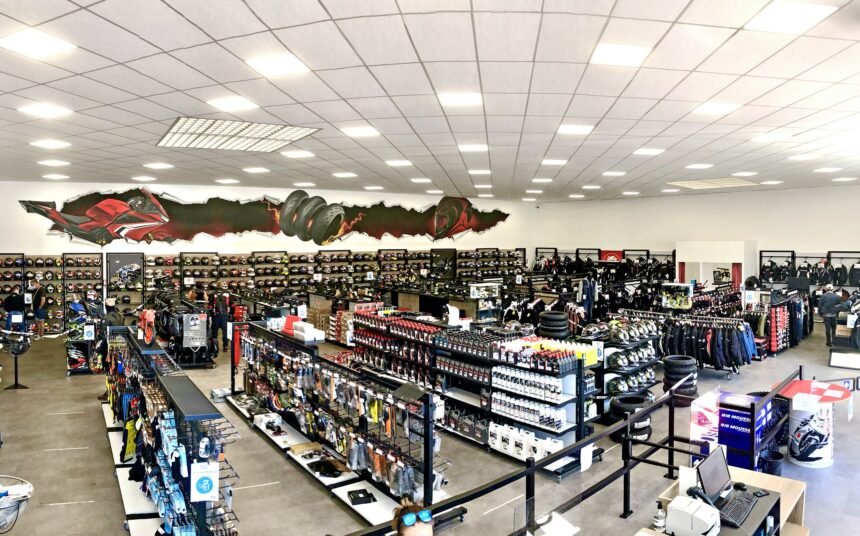 Vue panoramique d'un magasin d'équipement et d'accessoires pour motocycles présentant des casques, des vestes, des gants, des pièces détachées et des vêtements sur des étagères et des rayonnages sous un éclairage zénithal lumineux.
