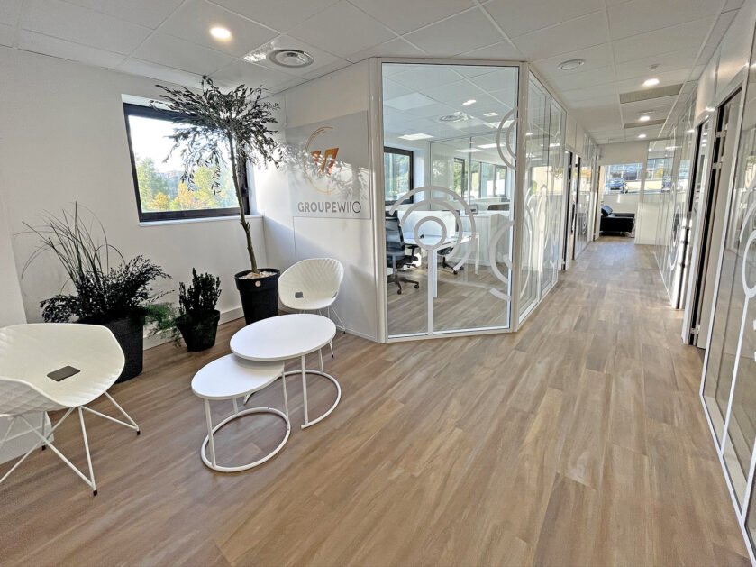 Hall de bureau moderne avec salles de réunion vitrées, chaises et tables blanches, plantes d'intérieur et sol en bois. La lumière naturelle pénètre par de grandes fenêtres.