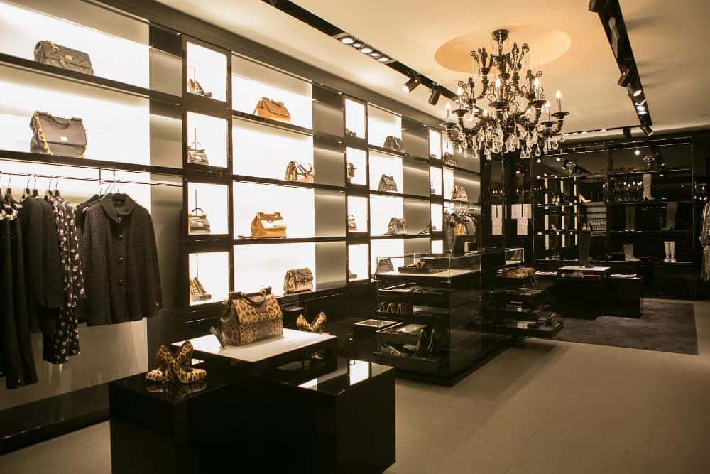 Boutique haut de gamme avec des étagères éclairées présentant des sacs à main, des vêtements sur des portants, un présentoir central en verre et un lustre au-dessus de la tête.
