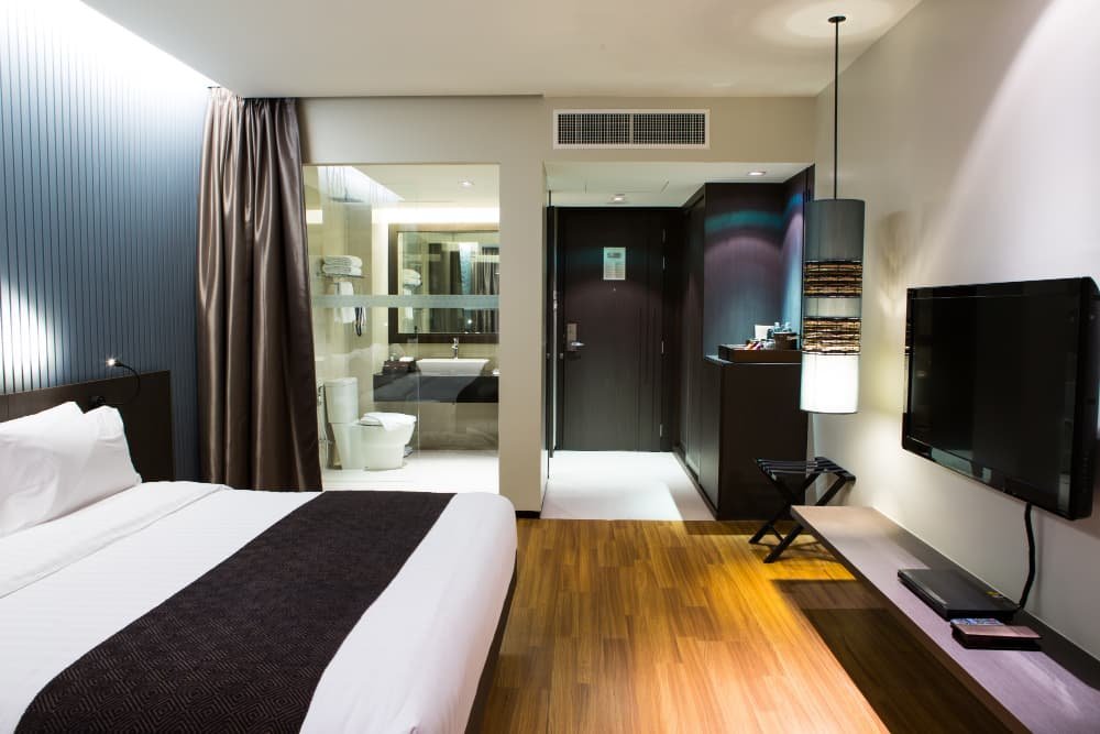Chambre d'hôtel moderne avec un lit bien fait, une télévision à écran plat, du parquet et une salle de bains ouverte avec des parois en verre.