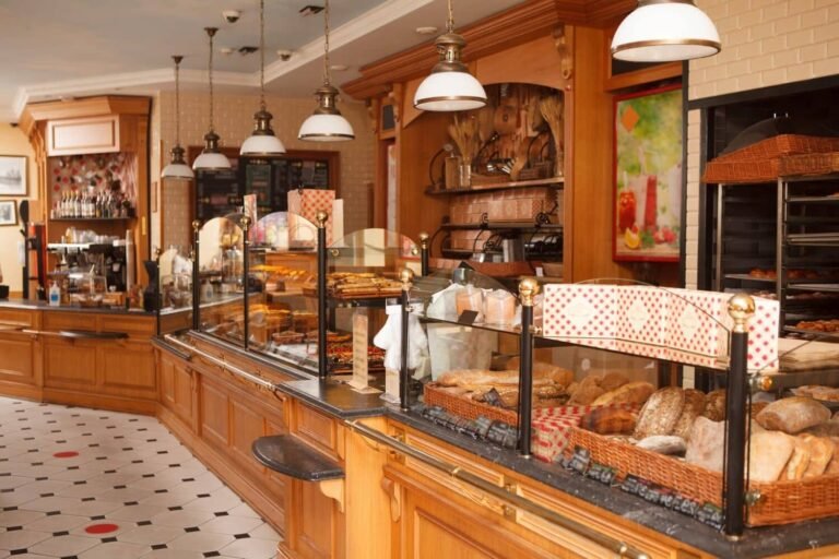Intérieur d'une boulangerie avec des comptoirs en bois présentant divers pains et pâtisseries derrière une vitre, avec des lampes suspendues et des étagères décoratives en arrière-plan.