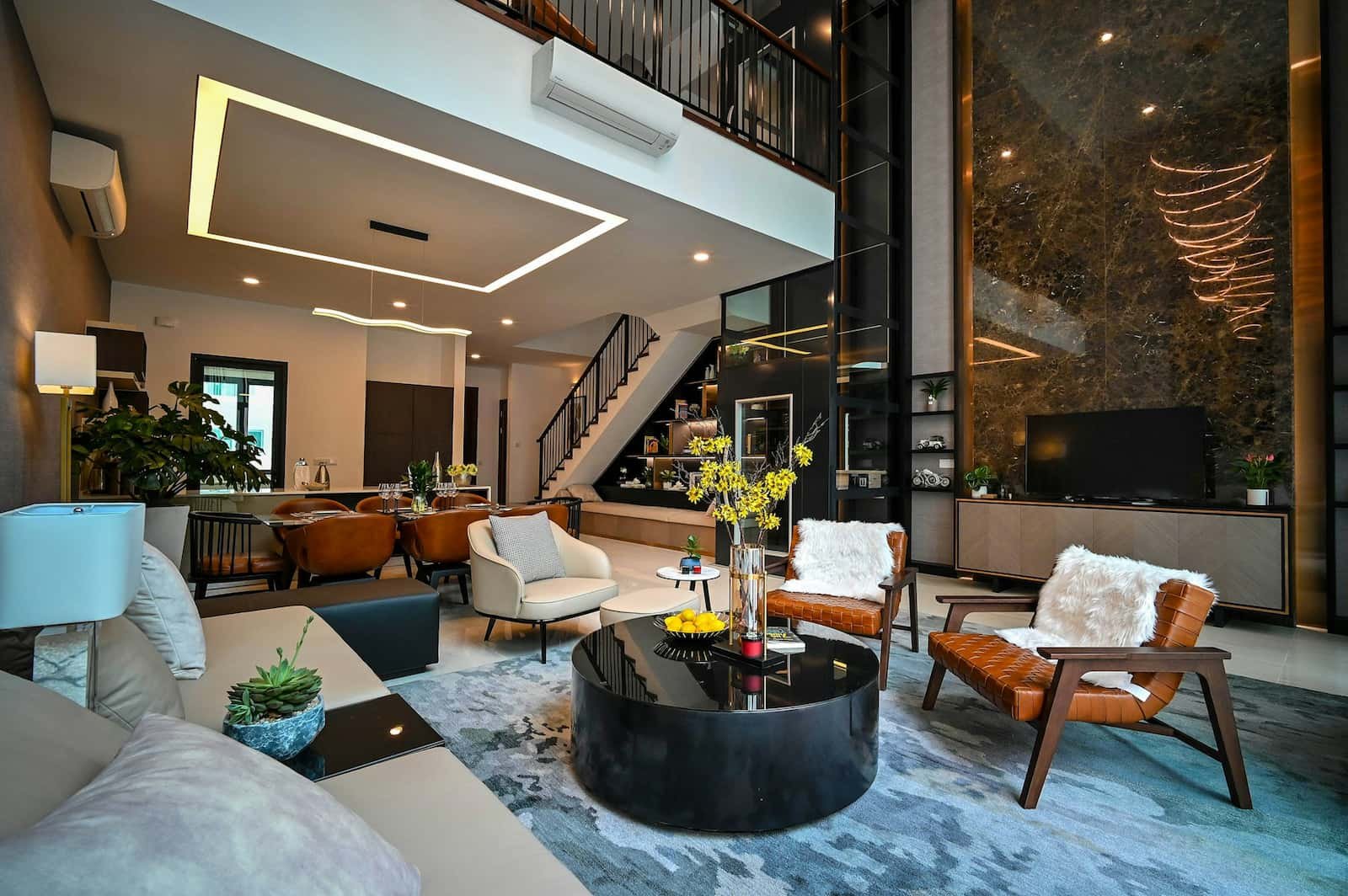 Salon moderne à aire ouverte avec mobilier contemporain, table basse en verre, plantes décoratives et escalier menant à l'étage supérieur. Tons neutres, éclairage d'appoint et grandes fenêtres.