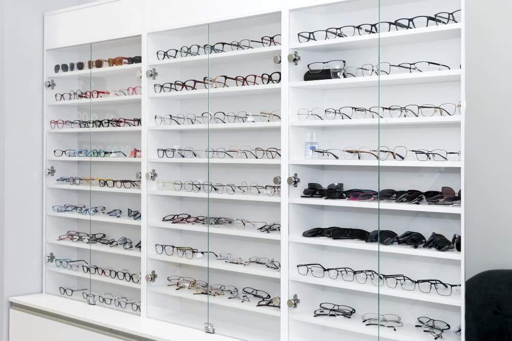 Une vitrine blanche avec des portes en verre montrant plusieurs rangées de lunettes de vue et de soleil assorties, disposées proprement sur des étagères.