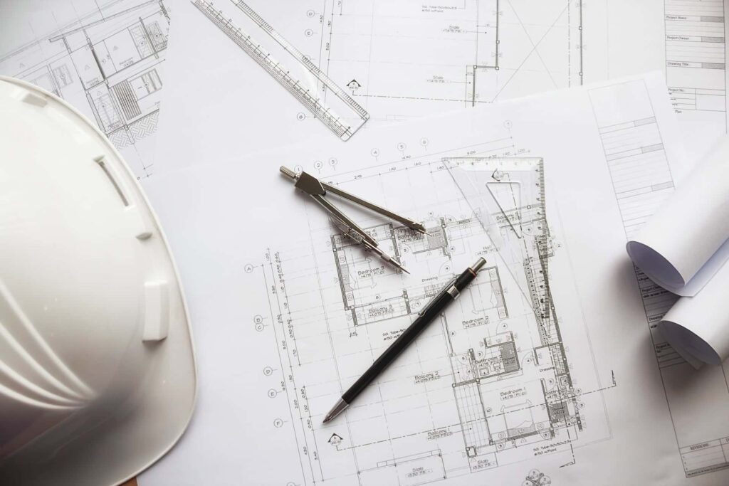 Des plans d'architecte, un casque blanc, un compas, un porte-mine, une règle et des plans enroulés sont disposés sur un bureau.