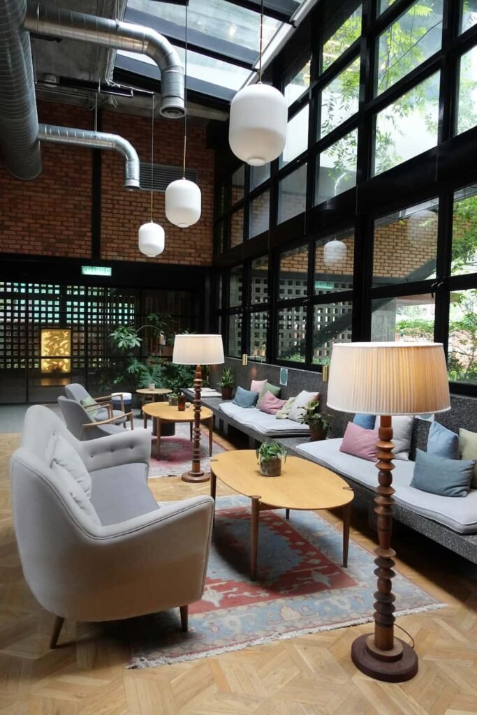 Salon moderne avec de grandes fenêtres, des murs en briques apparentes, des fauteuils gris, des canapés, des tables en bois, des plantes d'intérieur et des lampes globe suspendues.