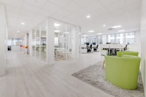 Espace de bureau moderne avec des salles de réunion en verre, des bureaux blancs, des chaises vertes et un tapis gris sur un sol en bois clair, avec de grandes fenêtres apportant de la lumière naturelle.
