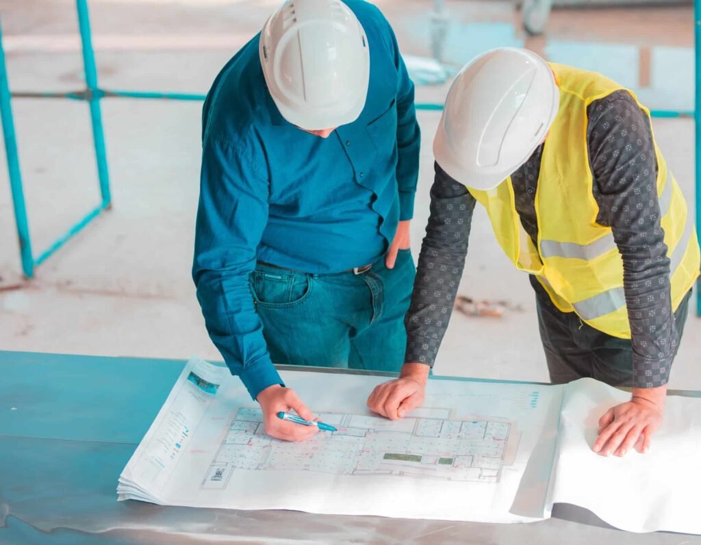 Deux personnes portant des casques de sécurité examinent des plans d'architecture sur une table sur un chantier de construction ; l'une pointe un détail avec un stylo tandis que l'autre regarde.