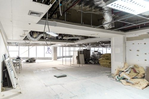 Intérieur d'un grand espace commercial inachevé avec des tuyaux de plafond exposés, des matériaux de construction éparpillés et des plaques de placoplâtre appuyées contre le mur.