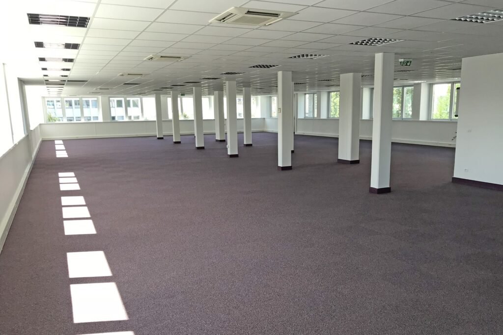 Grand espace de bureau vide avec une moquette violette, des murs blancs, de nombreuses colonnes de soutien et un réseau de plafonniers et d'unités de conditionnement d'air. La lumière du soleil pénètre par les fenêtres d'un côté.