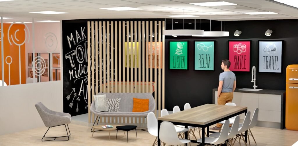 Salle de pause d'un bureau moderne avec une cloison en bois, des affiches colorées, un coin salon, une longue table avec des chaises, et une personne debout près d'un évier contre un mur d'accent noir.