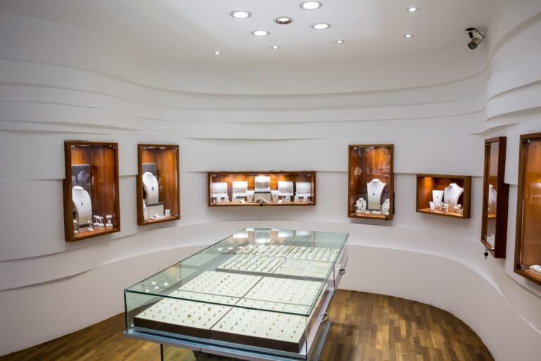 Intérieur d'une bijouterie avec des vitrines en verre présentant des bagues et des vitrines murales présentant des colliers et d'autres bijoux, sur un fond blanc et moderne.