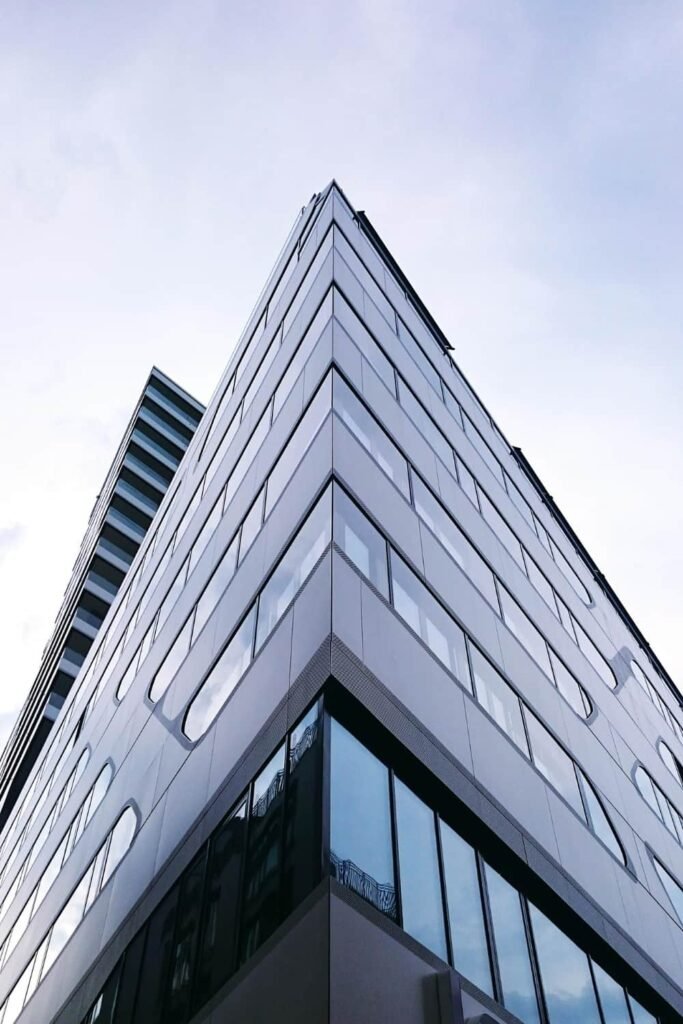 Un immeuble de bureaux moderne en verre et en métal, aux lignes anguleuses et aux fenêtres réfléchissantes, photographié depuis un angle bas et ascendant sur fond de ciel nuageux.