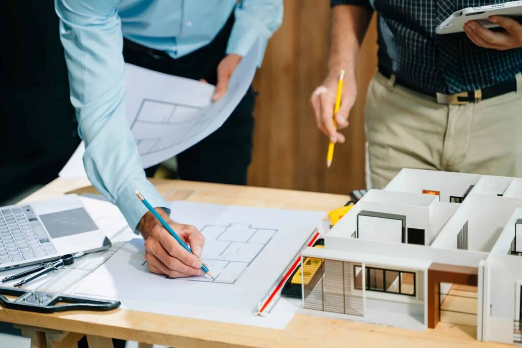 Deux personnes examinent et annotent des plans d'architecture sur un bureau où se trouvent également un ordinateur portable, une règle graduée et une maquette.