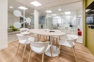 Salle de réunion de bureau moderne avec une table ronde en bois, six chaises blanches, des murs en verre et une télévision montée sur un mur d'accent vert.