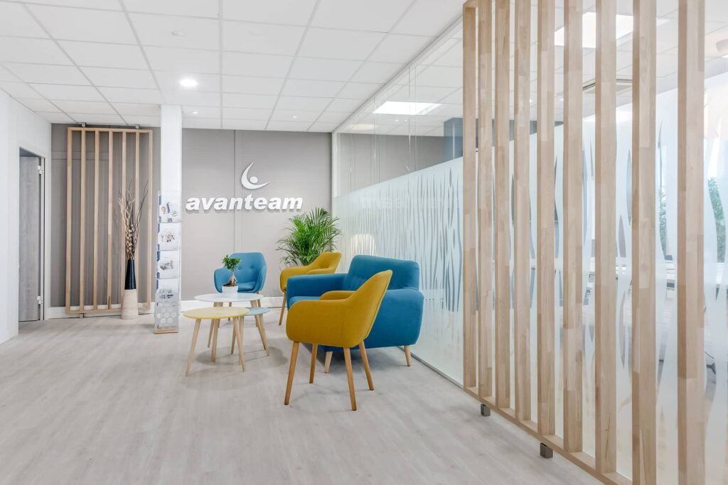 Salle d'attente de bureau moderne avec des chaises bleues et jaunes, des petites tables, des plantes décoratives et des murs en verre dépoli. Le logo "Avanteam" est affiché sur le mur du fond.
