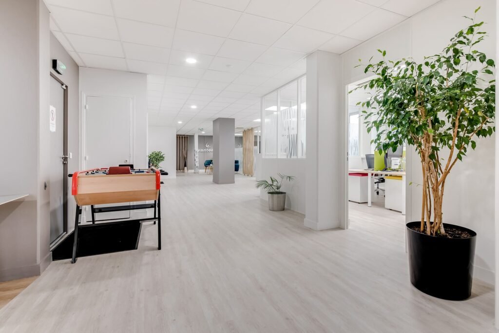 Couloir de bureau moderne avec un sol en bois clair, une plante en pot, un baby-foot et des portes ouvertes menant à des espaces de travail avec des bureaux et des chaises.
