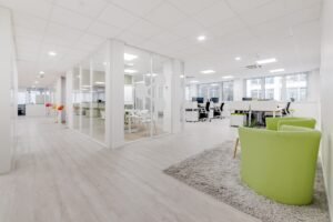 Bureau moderne à aire ouverte avec salle de réunion vitrée, bureaux blancs, chaises vertes sur un tapis gris, et lumière naturelle provenant de grandes fenêtres.