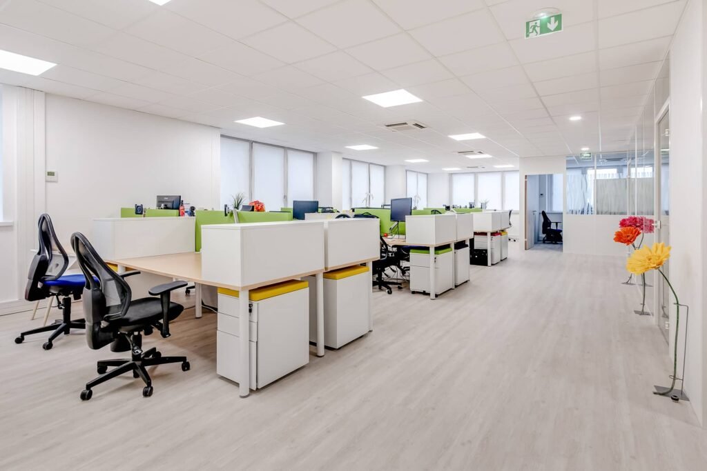 Bureau moderne à aire ouverte avec des bureaux blancs, des chaises noires, des écrans de bureau et des cloisons vertes. De grandes fenêtres et un sol en bois clair créent un environnement lumineux et spacieux.