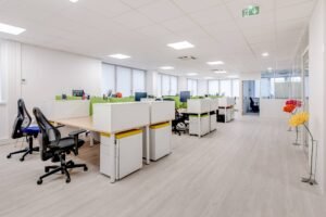 Bureau moderne à aire ouverte avec des bureaux blancs, des chaises noires, des écrans de bureau et des cloisons vertes. De grandes fenêtres et un sol en bois clair créent un environnement lumineux et spacieux.
