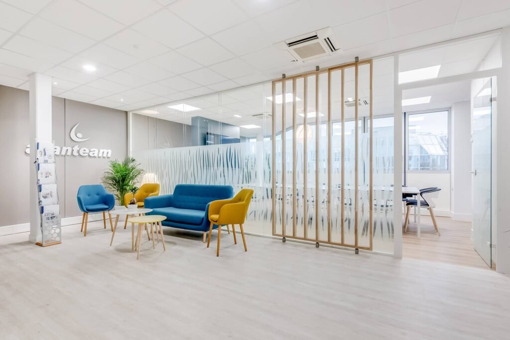 Salle d'attente d'un bureau moderne avec des chaises bleues et jaunes, une petite table, une plante en pot et une cloison en verre dépoli à côté d'une salle de conférence.