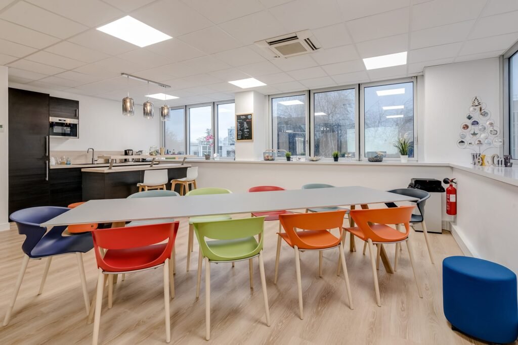 Une salle de pause moderne avec une longue table, des chaises colorées et une kitchenette.