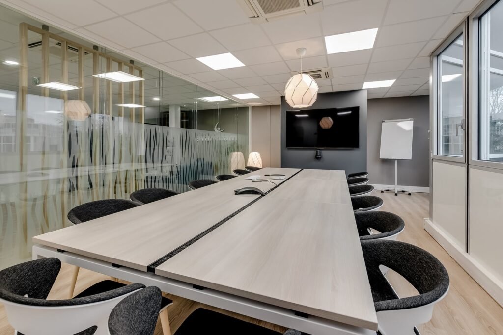 Salle de conférence moderne équipée d'une longue table, de chaises noires, d'un écran mural, d'un tableau à feuilles mobiles, d'une cloison en verre et de grandes fenêtres laissant entrer la lumière naturelle.