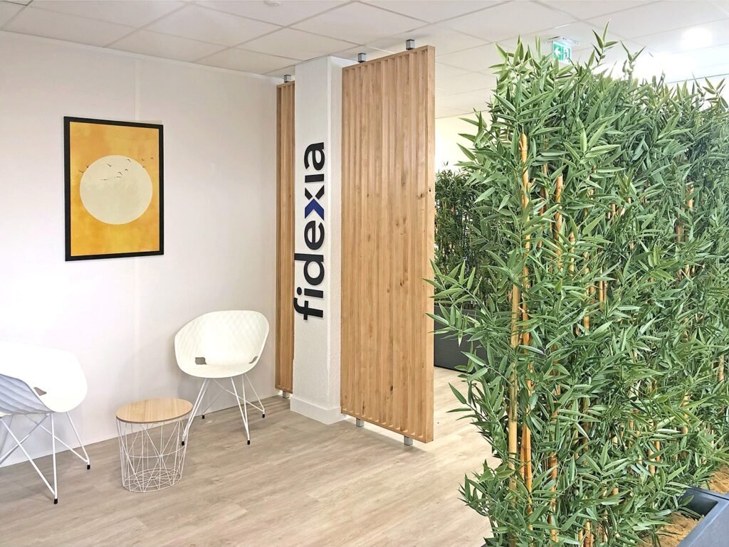 Salle d'attente du bureau avec deux chaises blanches, une petite table, une œuvre d'art encadrée, une cloison avec le logo "fidexia" et une grande plante artificielle en bambou.