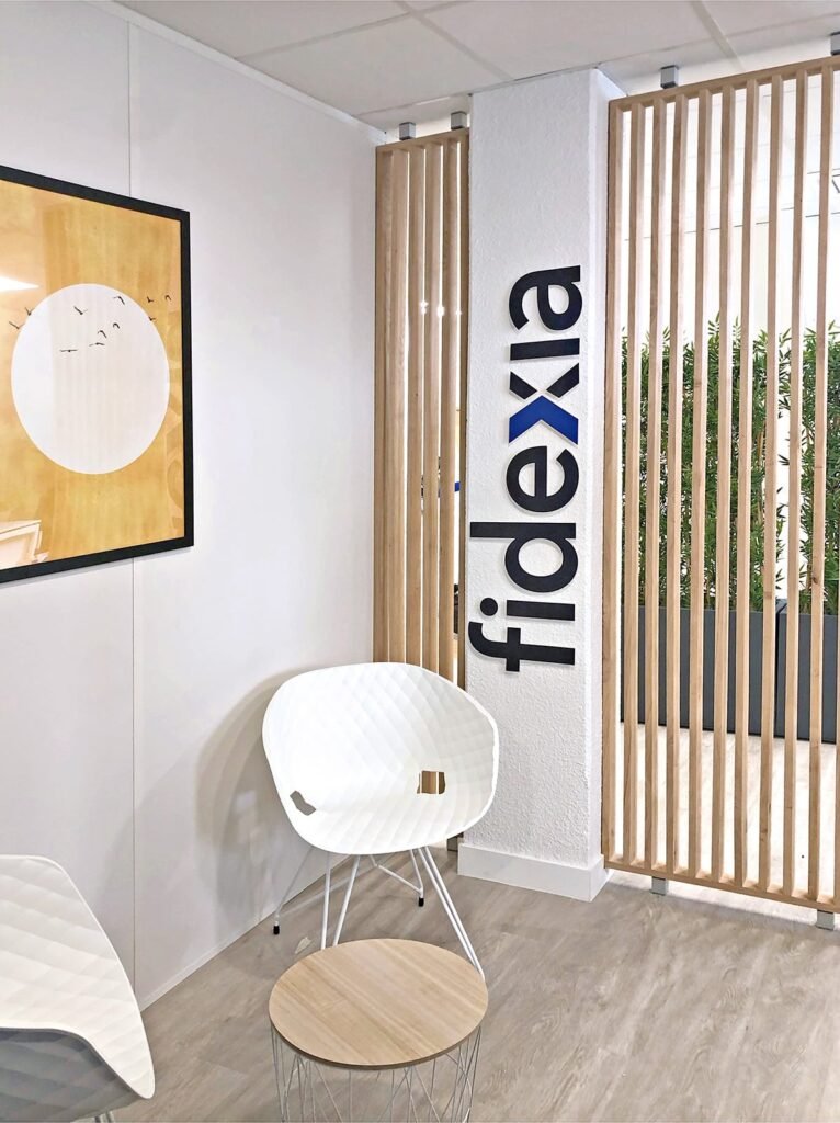 Salle d'attente d'un bureau moderne avec des chaises blanches, une petite table ronde, une cloison en lattes de bois, des œuvres d'art murales et le mot "fidxia" affiché verticalement sur le mur.