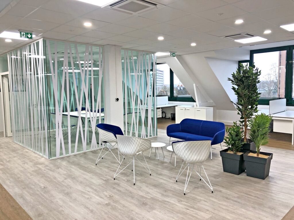 Espace de bureau moderne avec salle de réunion vitrée, canapé bleu, chaises blanches, petites tables, plantes en pot et grandes fenêtres offrant une lumière naturelle.