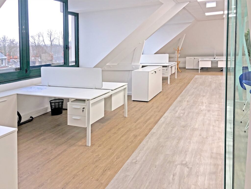 Espace de bureau moderne avec des bureaux, des chaises et des armoires blancs disposés le long de grandes fenêtres, un sol en bois clair et un mur en verre sur le côté droit.