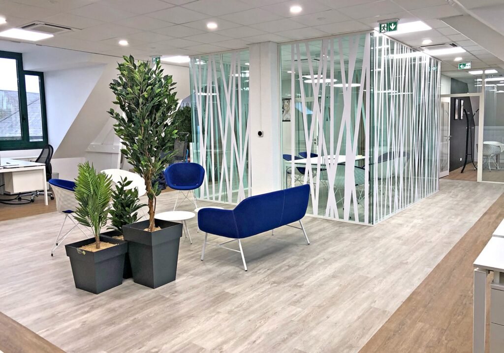 Espace de bureau moderne avec salle de réunion vitrée, sièges bleus, plantes en pot et bureaux dans un cadre lumineux et minimaliste avec lumière naturelle et sol en bois.