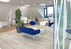 Bureau moderne avec des bureaux blancs, des chaises noires, un petit coin salon avec des sièges bleus et blancs, une table ronde, une plante en pot et une grande fenêtre qui apporte de la lumière naturelle.