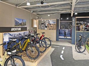 Une salle d'exposition présente divers vélos sur des supports, avec des panneaux indiquant "Aventure", "Loisirs" et "Atelier". De grandes photos de scènes extérieures ornent les murs.