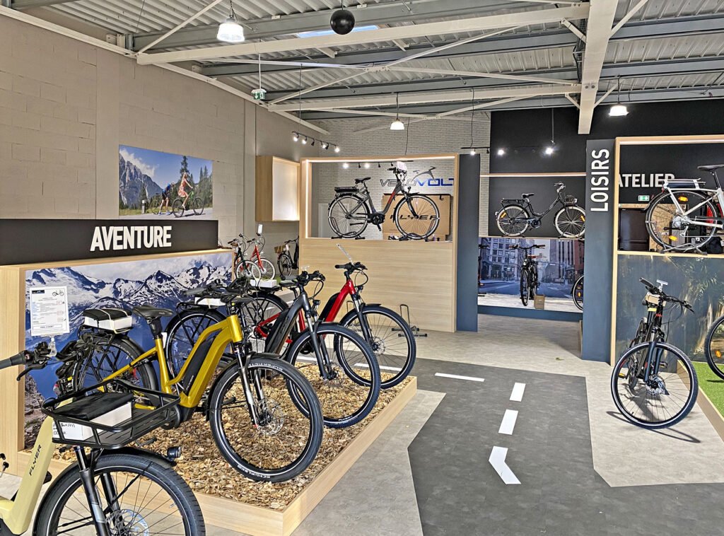 L'intérieur d'un magasin de vélos moderne présente divers vélos sur des supports, avec des panneaux étiquetés "AVENTURE", "LOISIRS" et "ATELIER", et des affiches sur le thème du cyclisme sur les murs.