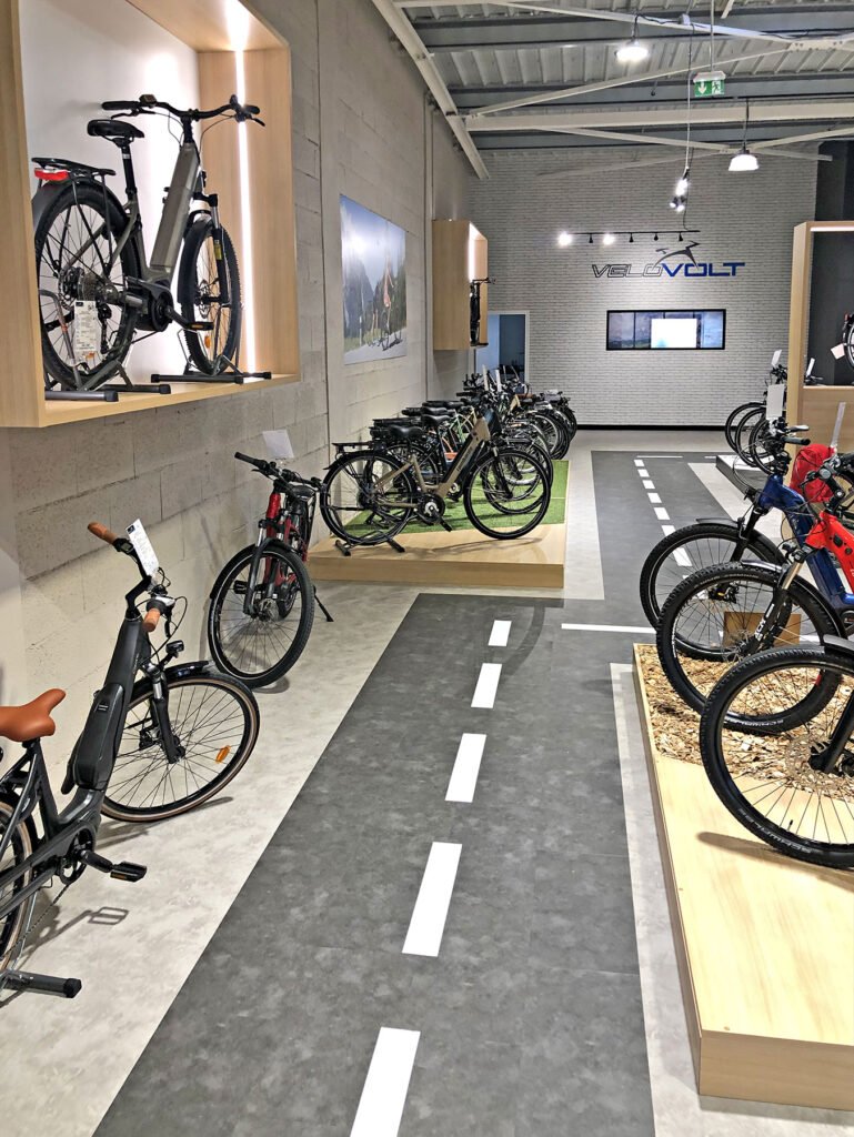 Salle d'exposition de bicyclettes avec divers vélos présentés sur des supports et des étagères, avec un sol à thème routier sous les vélos.