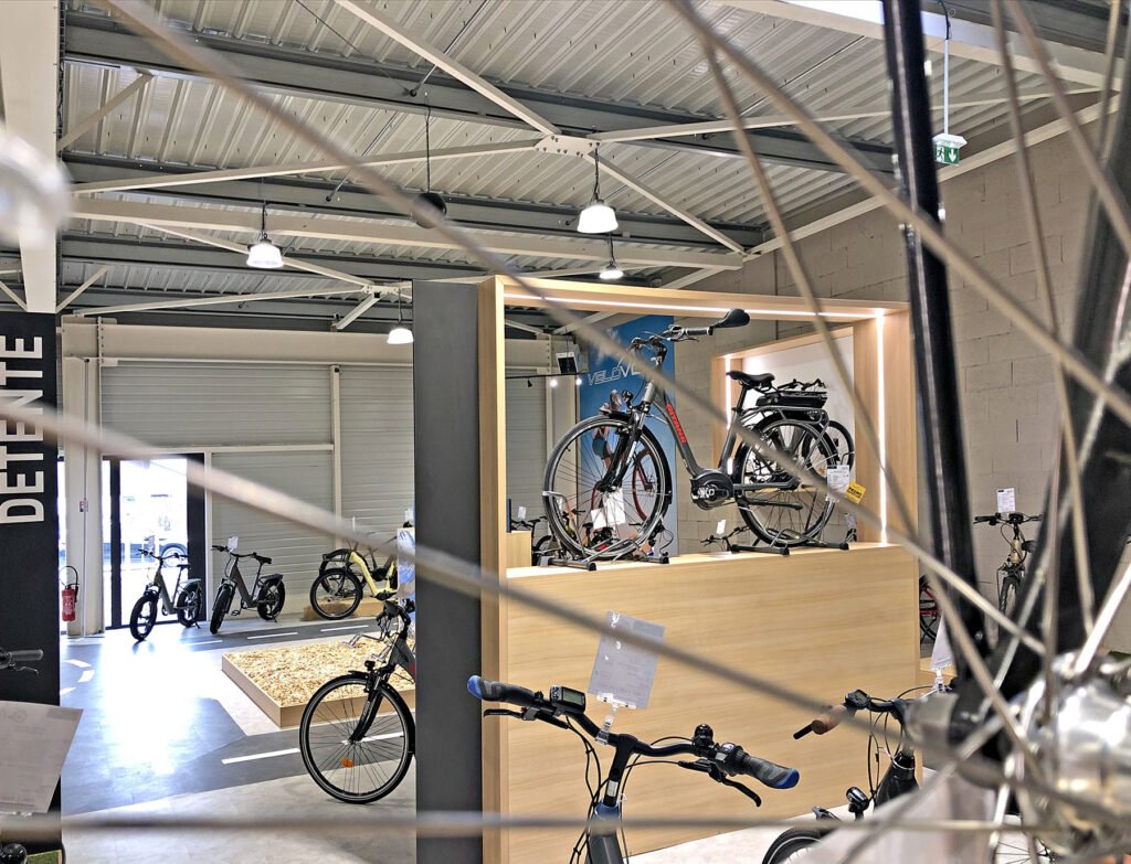 Une salle d'exposition de bicyclettes moderne présente divers vélos sur le sol et sur une plate-forme en bois, avec un plafond métallique, de grandes lumières et des roues de bicyclettes visibles au premier plan.