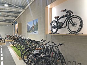 Une rangée de vélos est exposée dans une salle d'exposition, avec un vélo électrique monté sur une étagère en bois au-dessus et une peinture sur le thème du cyclisme accrochée au mur.