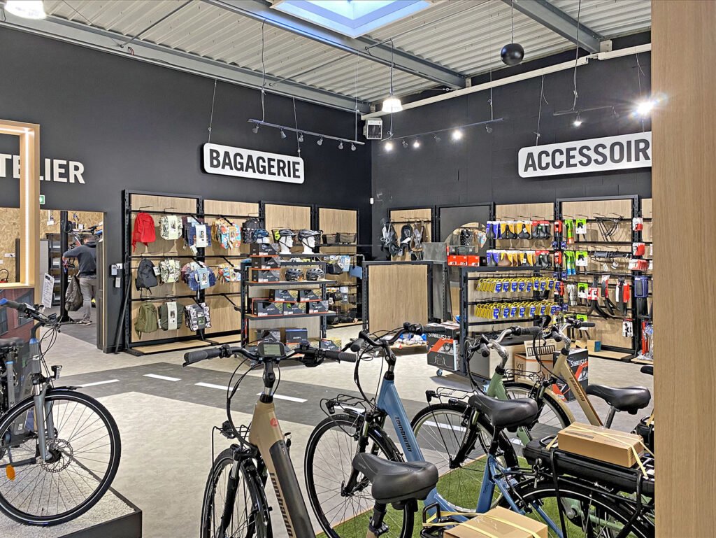 Intérieur d'un magasin de vélos avec des vélos au premier plan et des présentoirs de sacs et d'accessoires sur les murs sous les enseignes "BAGAGERIE" et "ACCESSOIRE".