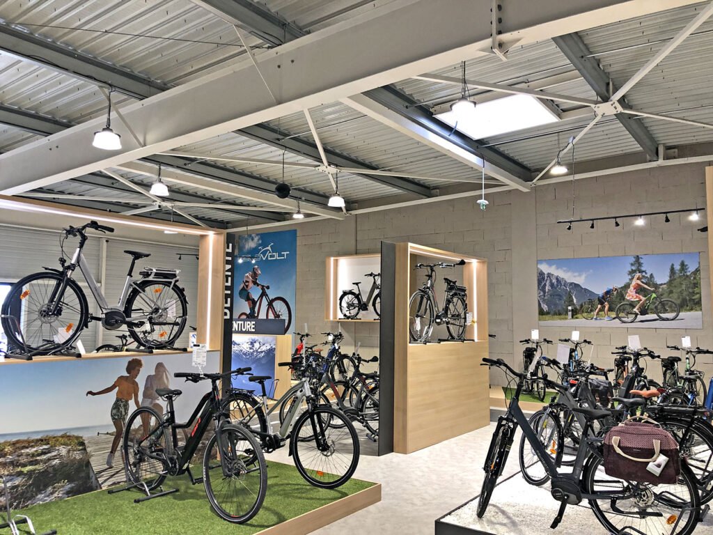 Salle d'exposition de bicyclettes avec des vélos électriques exposés sur des supports et des plates-formes, des affiches de cyclistes sur les murs et un éclairage d'intérieur au plafond.