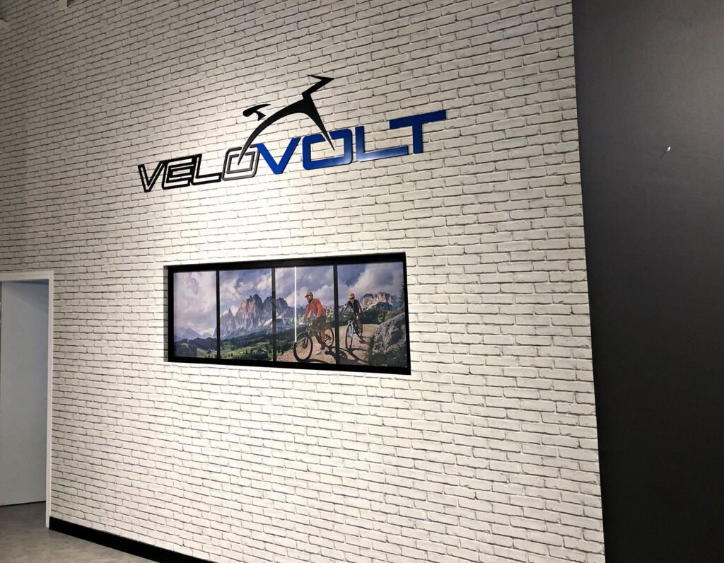 Mur de briques blanches avec le logo "VELOVOLT" et un cycliste stylisé au-dessus d'une photo encadrée montrant des cyclistes sur une piste de montagne.