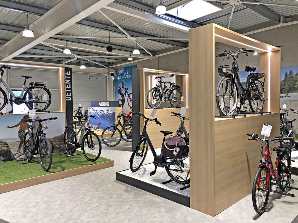 Les vélos sont exposés sur des plates-formes surélevées et sur le sol dans une salle d'exposition bien éclairée, avec des installations modernes en bois et des affiches sur le cyclisme sur les murs.