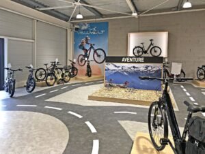 Salle d'exposition intérieure de bicyclettes présentant divers vélos sur des stands, avec des marquages routiers sur le sol et des affiches sur le thème de l'aventure sur les murs.