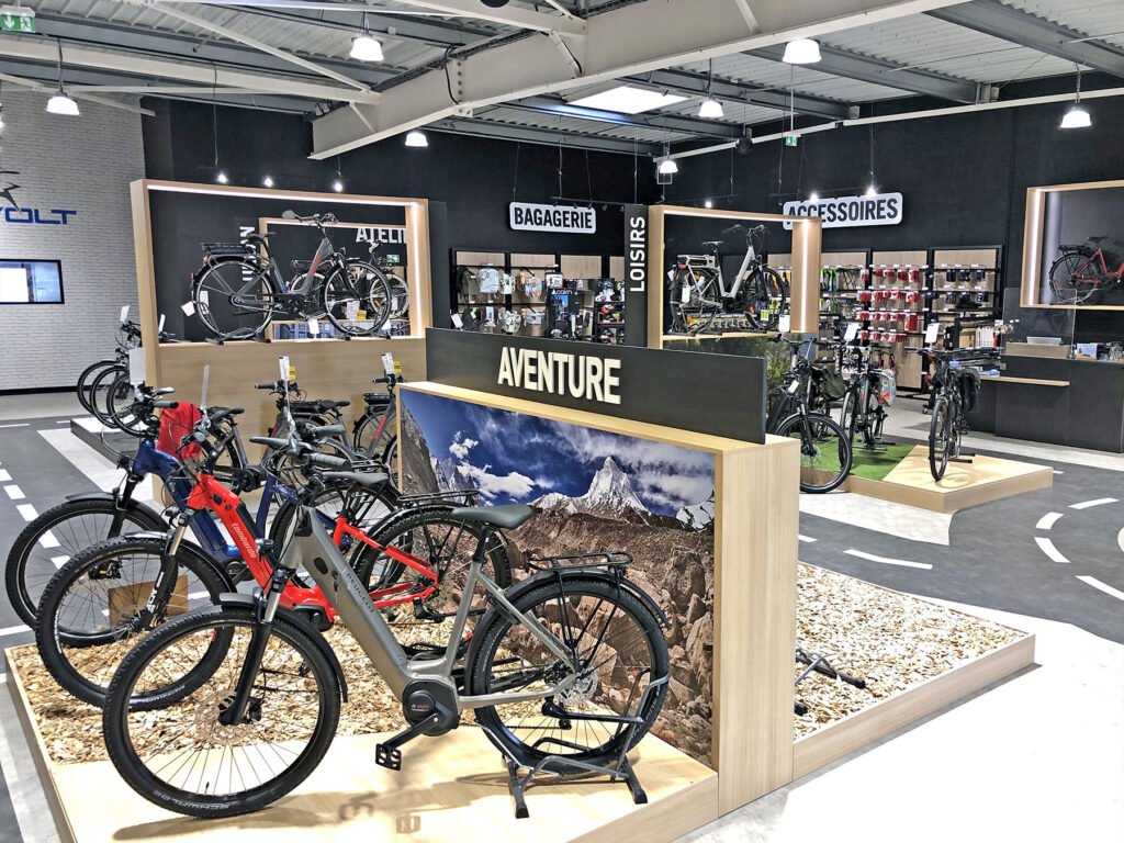 Intérieur d'un magasin de bicyclettes avec des vélos de montagne en exposition, des supports en bois étiquetés "Aventure" et divers accessoires de cyclisme sur des étagères à l'arrière-plan.