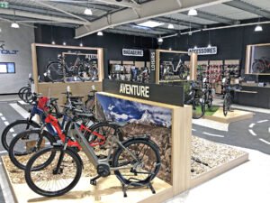 L'intérieur d'un magasin de vélos moderne présente divers vélos, accessoires et équipements sur des racks et des étagères, avec des panneaux en français indiquant les différentes sections.