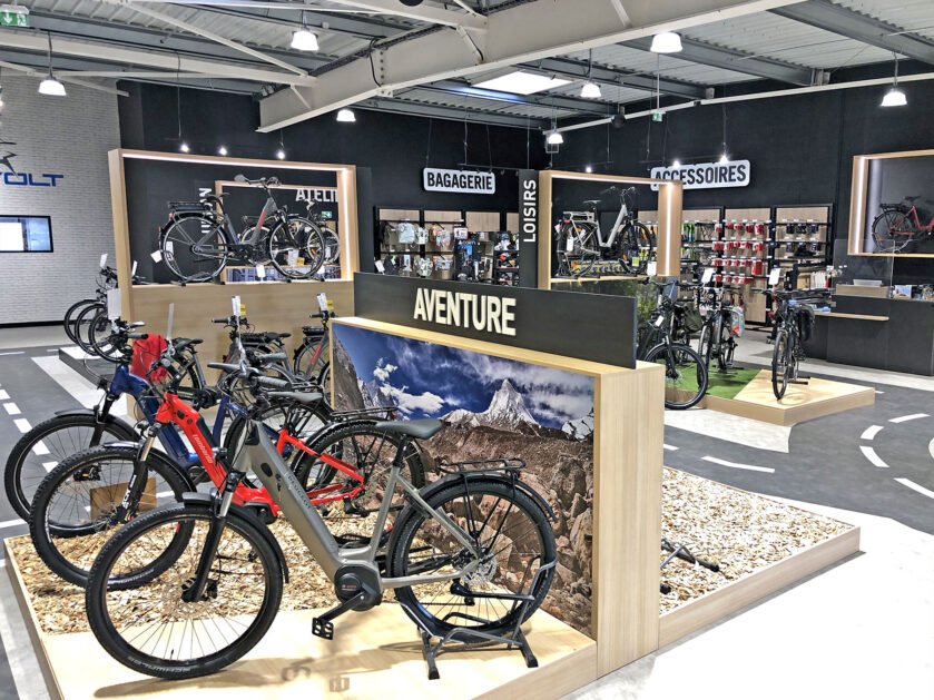 L'intérieur d'un magasin de vélos moderne présente divers vélos, accessoires et équipements sur des racks et des étagères, avec des panneaux en français indiquant les différentes sections.