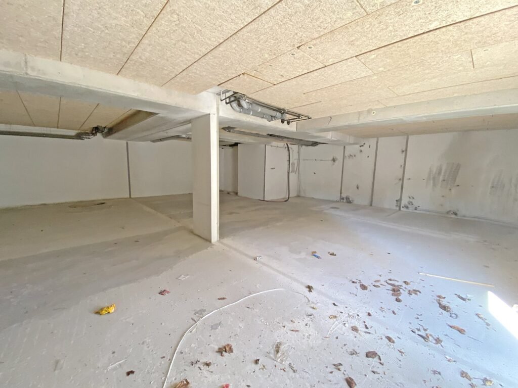 Une pièce non finie avec un sol et des murs en béton, des tuyaux apparents et un plafond en bois.