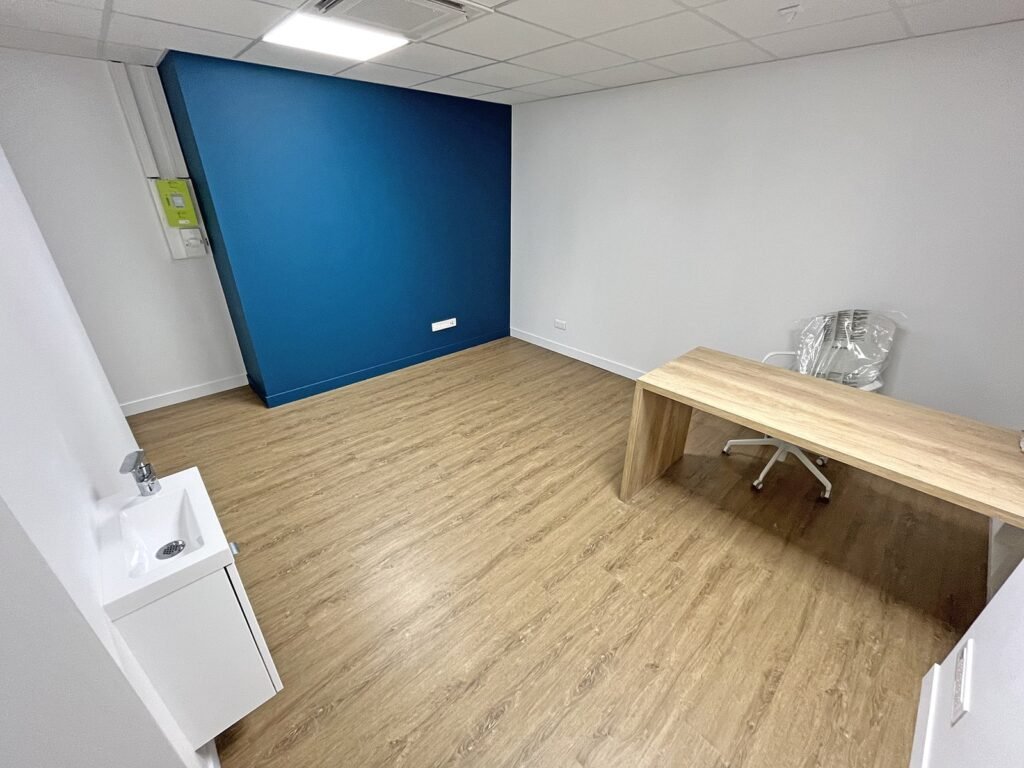 Bureau vide avec un sol en bois, un mur d'accent bleu, un petit évier blanc et un bureau en bois avec une chaise de bureau enveloppée.