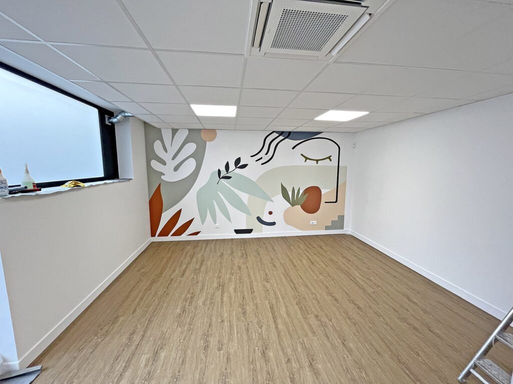 Une pièce moderne et vide avec un sol en bois, des dalles de plafond blanches et un mur orné d'une peinture murale abstraite colorée avec des formes géométriques et végétales.